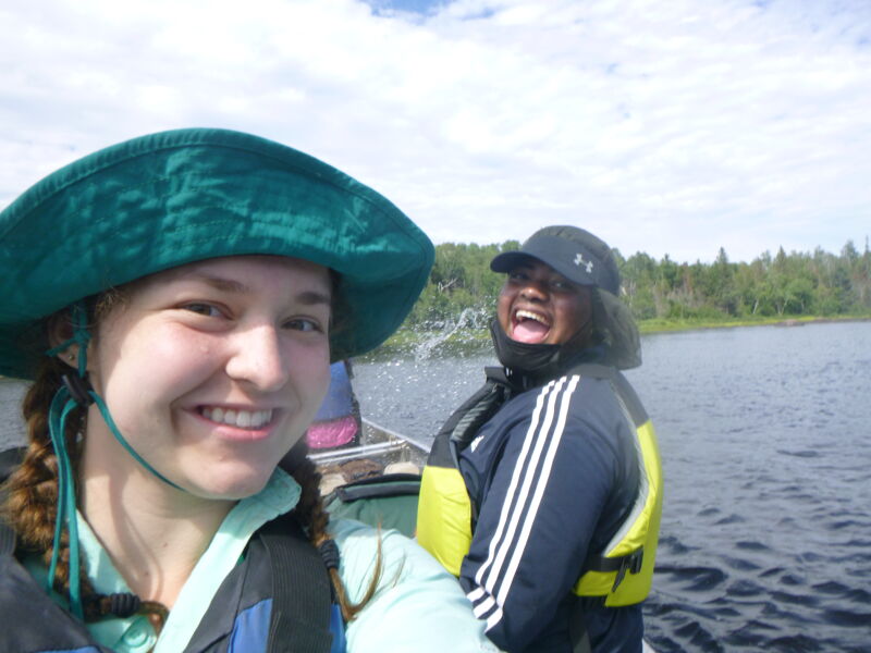 Two people are in a boat on a lake. The person in the foreground is wearing a green hat and smiling. The person in the background is wearing a black hat and a life jacket and is laughing. The lake is surrounded by trees. The sky is cloudy.